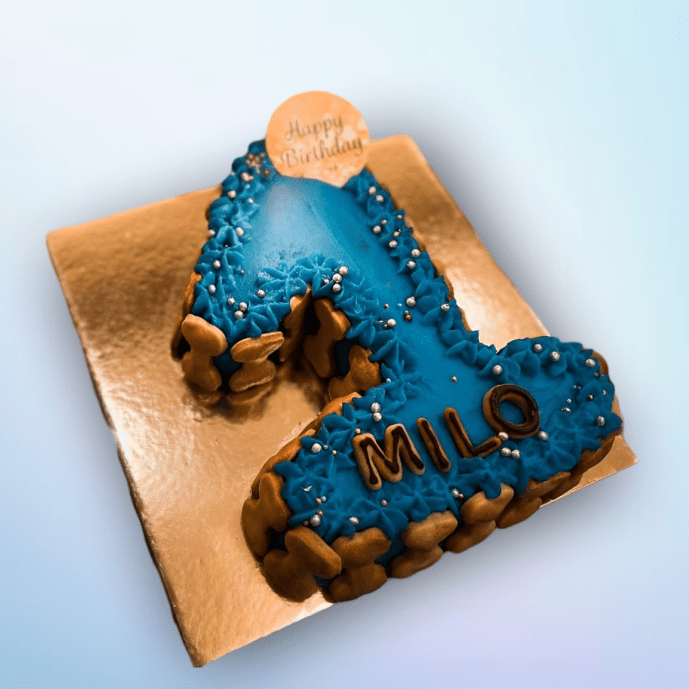 Blue number 1 dog birthday cake with name “Milo,” decorated with dog biscuits, frosting, and pearl