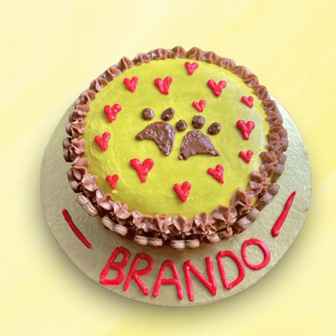 Round yellow dog cake with brown paw prints and red hearts, decorated with chocolate frosting and personalized name
