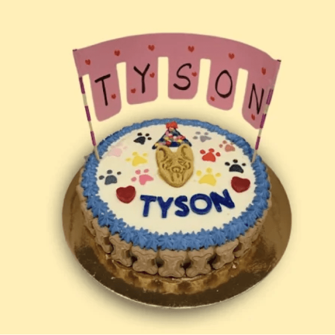 Round white dog birthday cake with blue border, colorful paw prints, and a custom Tyson banner, topped with a party hat biscuit