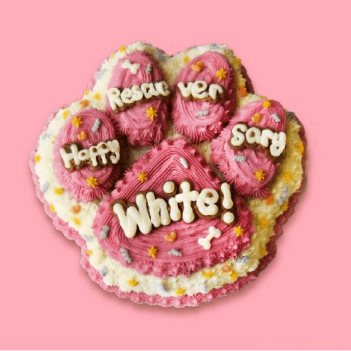 Pink and white paw-shaped dog birthday cake with a message and colorful star sprinkles