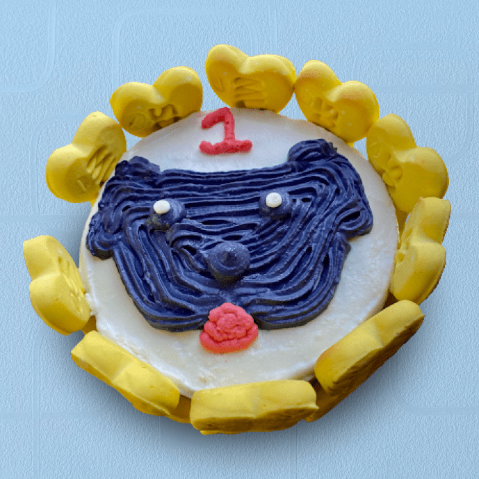 Dog-themed birthday cake with a cartoon face design, yellow heart-shaped biscuits around the edge, and a red “1” topper for a pup’s first birthday.
