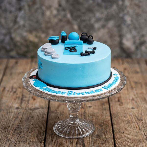 Light blue cake decorated with miniature fondant gym gear including a yoga mat and weights.