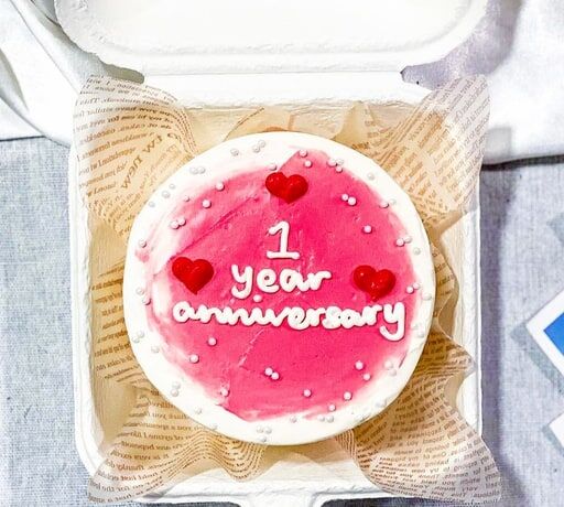 A pink cake with '1 year anniversary' written on it, with small red hearts and white dots, in a box