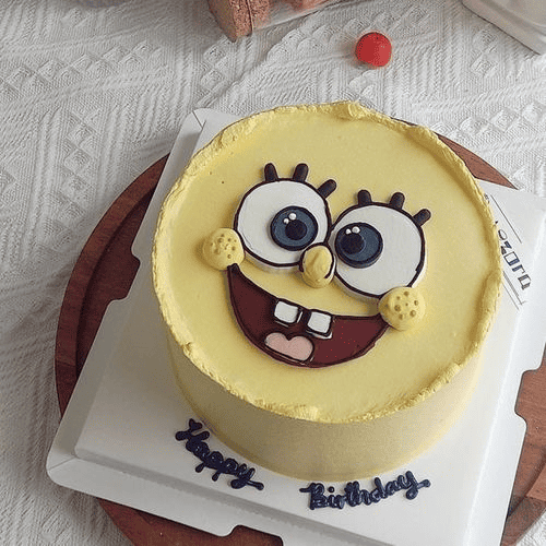 A yellow spongebob cartoon character-themed birthday cake with big eyes and a happy expression, sitting on a white cake board with 'Happy Birthday' written in blue.